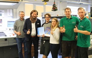 Team mit Büchern vor Druckmaschine, alle zeigen Daumen hoch und lächeln in die Kamera