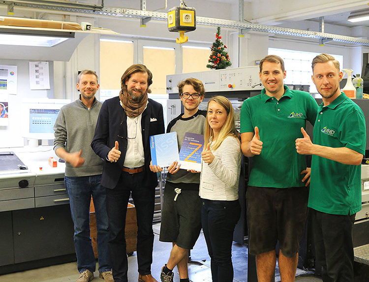 Team mit Büchern vor Druckmaschine, alle zeigen Daumen hoch und lächeln in die Kamera