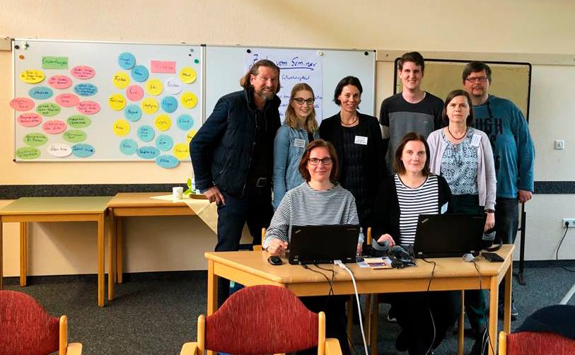 Seminargruppe vor Whiteboard, zwei Frauen sitzen an Laptops, alle schauen in die Kamera