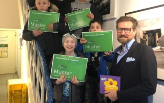 Kinder mit grünen MHD-Schildern auf Treppe, Mann hält Schokolade als Dank