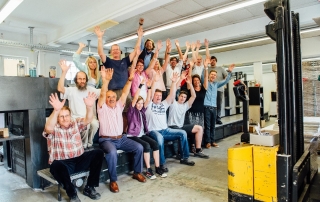 Gruppenfoto in der Druckerei: Das gesamte MHD-Team hebt fröhlich die Hände