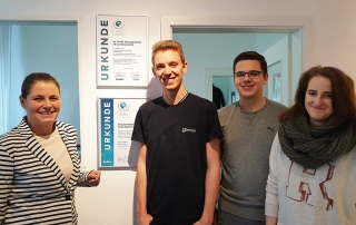 Vier junge Erwachsene stehen lächelnd vor zwei Urkunden an der Wand