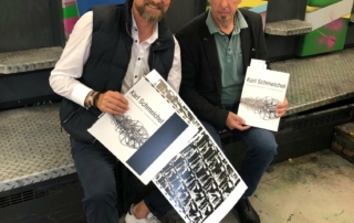 Zwei Männer halten Buch und Druckbogen mit dem Titel „Karl Schmeichel“ vor Druckmaschine