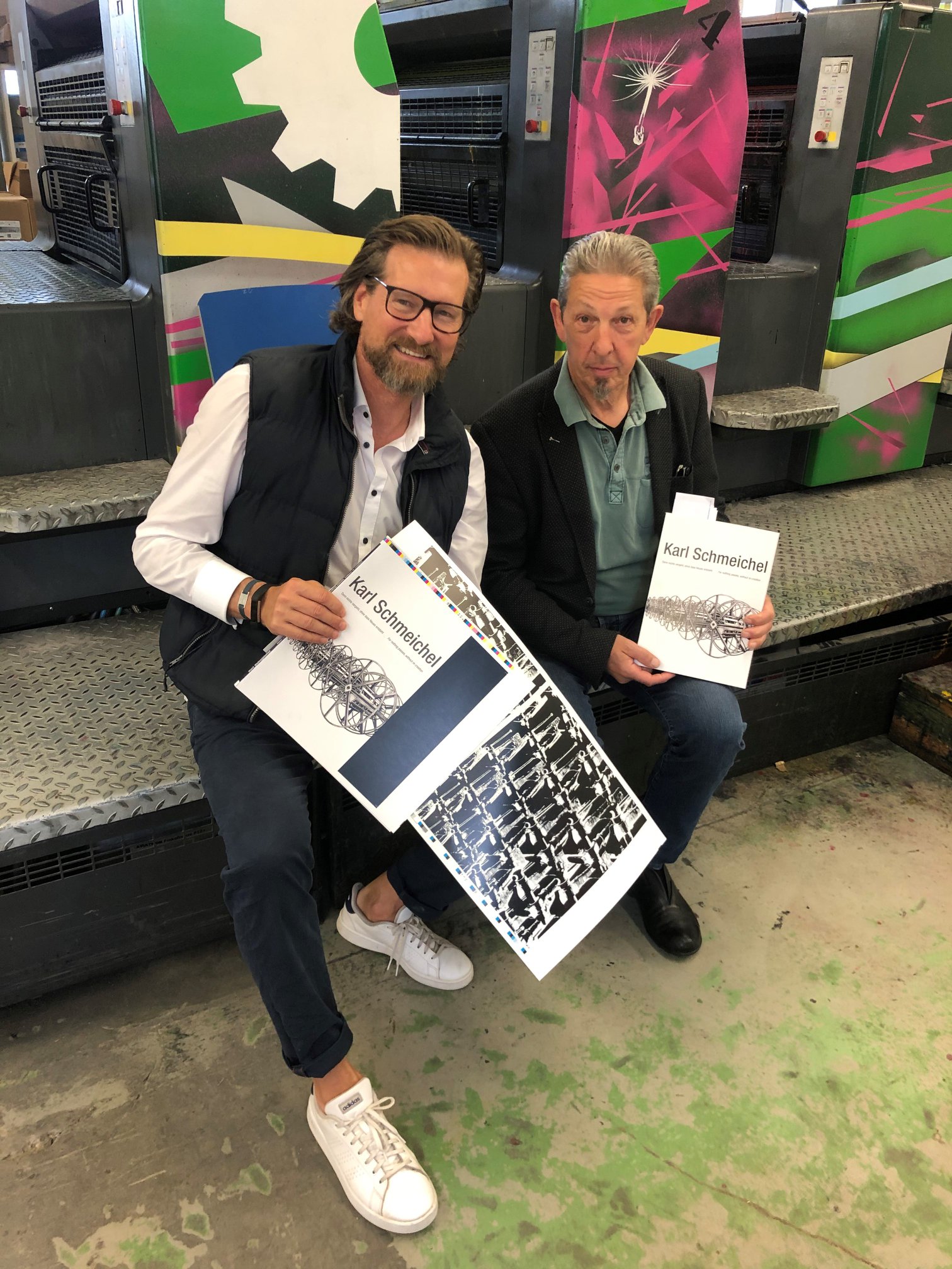 Zwei Männer halten Buch und Druckbogen mit dem Titel „Karl Schmeichel“ vor Druckmaschine