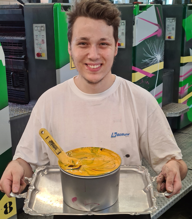 Junger Mann mit Druckfarbe in Dose auf Tablett vor bunter Offsetdruckmaschine