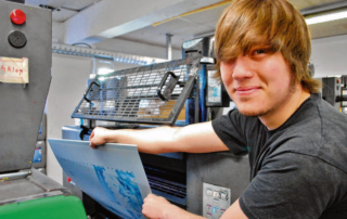 Junger Mann bedient Druckmaschine und hält eine Druckplatte in der Hand
