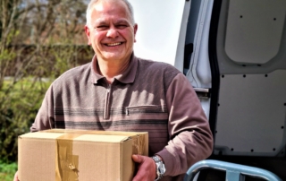Mann mit grauen Haaren und Paket vor Fahrzeug, freundlich lächelnd bei Sonnenschein