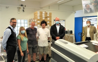 Sechs Personen mit Masken posieren in einer Druckerei, Kartons im Hintergrund