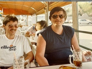 Zwei Männer mit Sonnenbrillen sitzen im Straßencafé, trinken Cola, 80er- oder 90er-Jahre-Stil