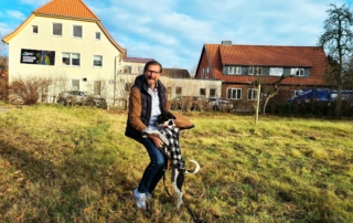 Mann steht mit Hund auf Wiese vor Gebäude mit Schild „MHD MedienHaus Druckerei“
