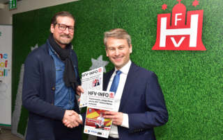 Zwei Männer halten HFV-Infoflyer und schütteln sich vor grünem Hintergrund die Hand