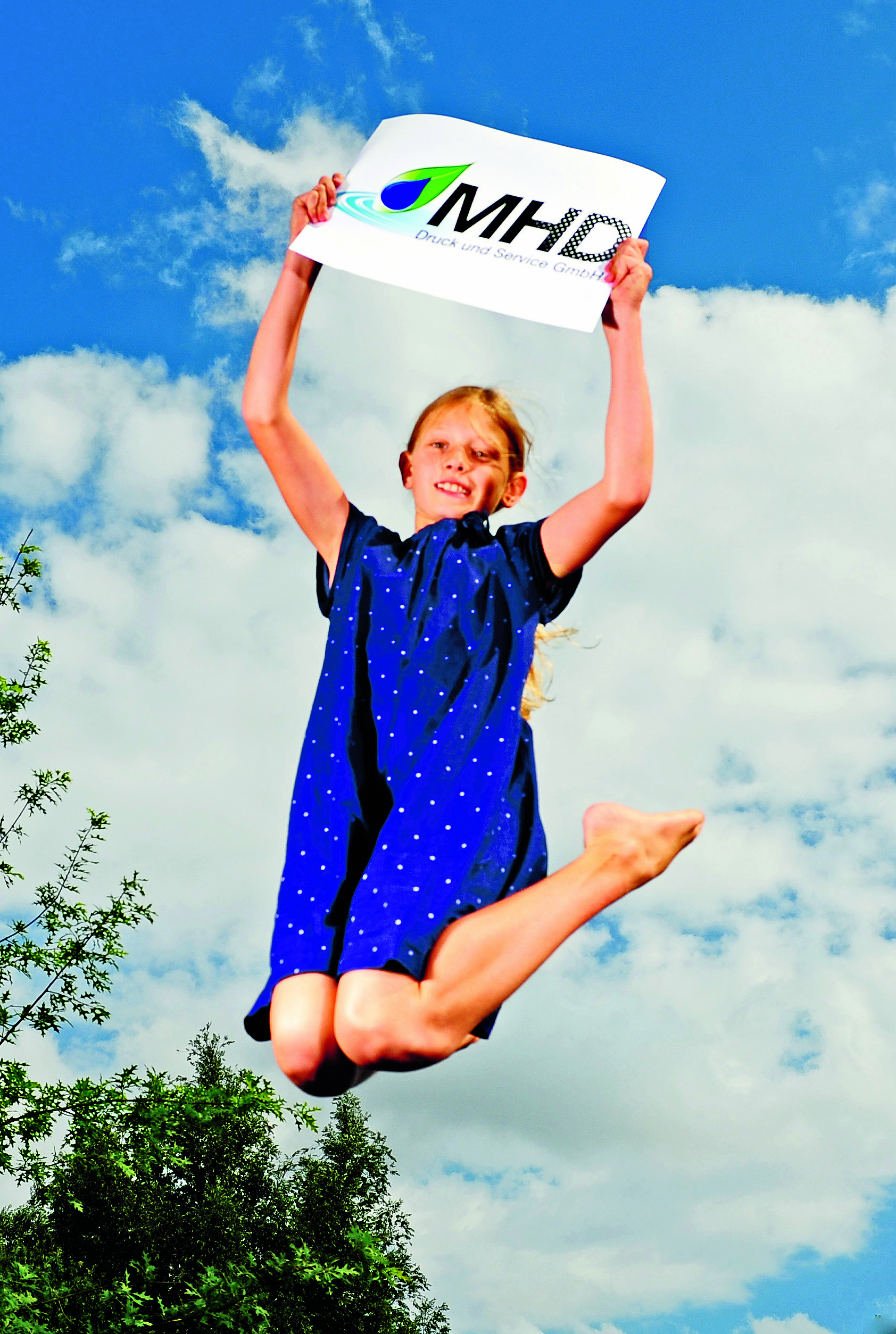 Mädchen springt fröhlich in die Luft und hält ein Schild mit MHD-Logo vor blauem Himmel