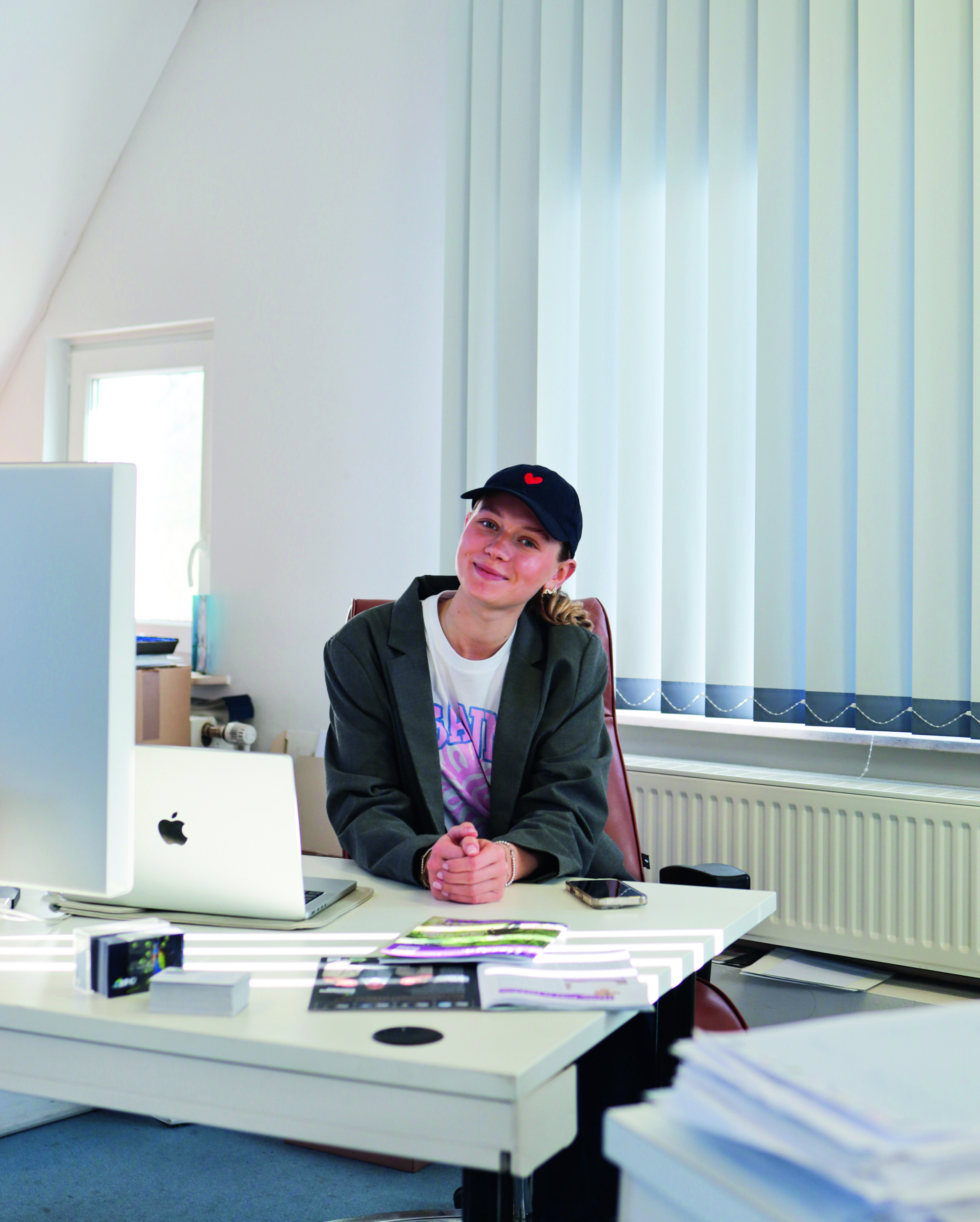 Finja_Heute Frau mit schwarzer Kappe sitzt lächelnd an einem Schreibtisch mit Computer und Unterlagen